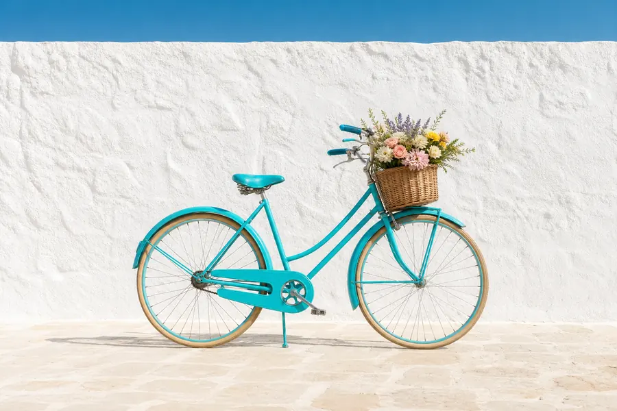 A turquoise bicycle with a flower basket against a sunlit white wall