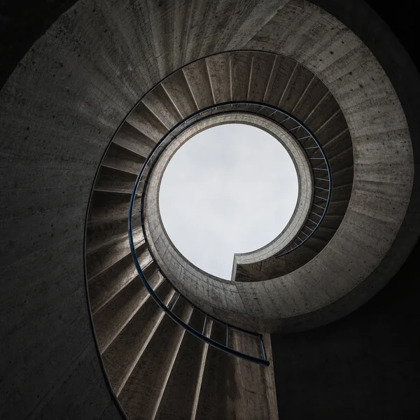 A spiral concrete staircase seen from below, opening onto an oval of white sky.