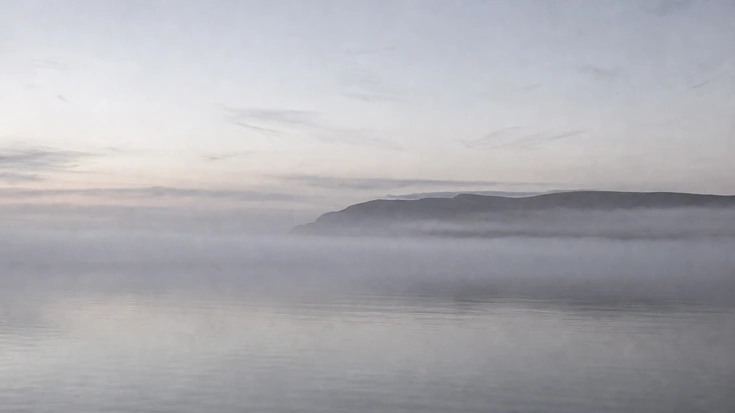 A sheet of mist lying on still water, a headland barely visible on the horizon.