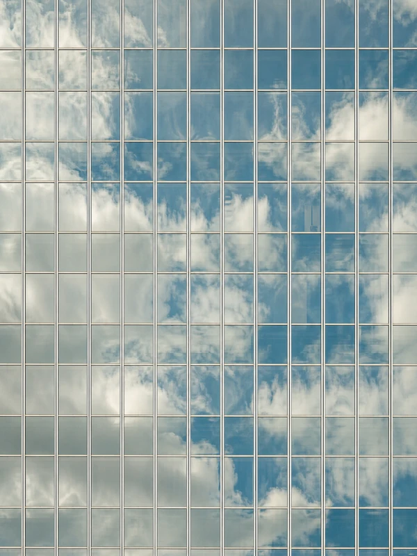 The glass façade of a tower, its grid of panes reflecting a blue sky and white clouds.