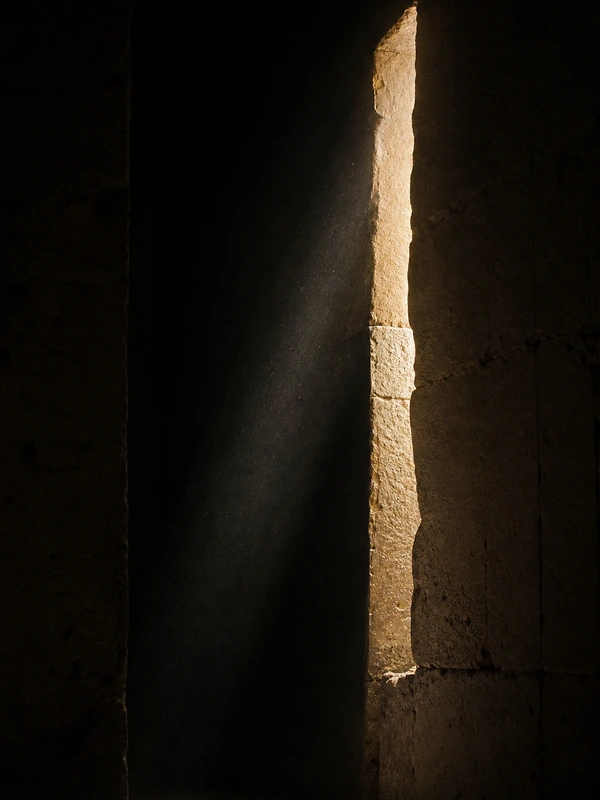 A shaft of light crossing a dark stone interior and striking the edge of a limestone wall.