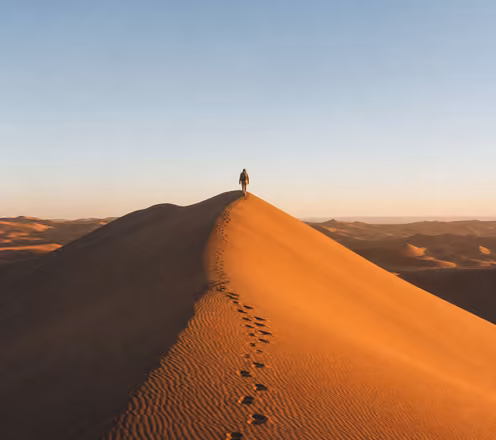 Person walking alone on a ridge of large sand dunes under clear blue sky.