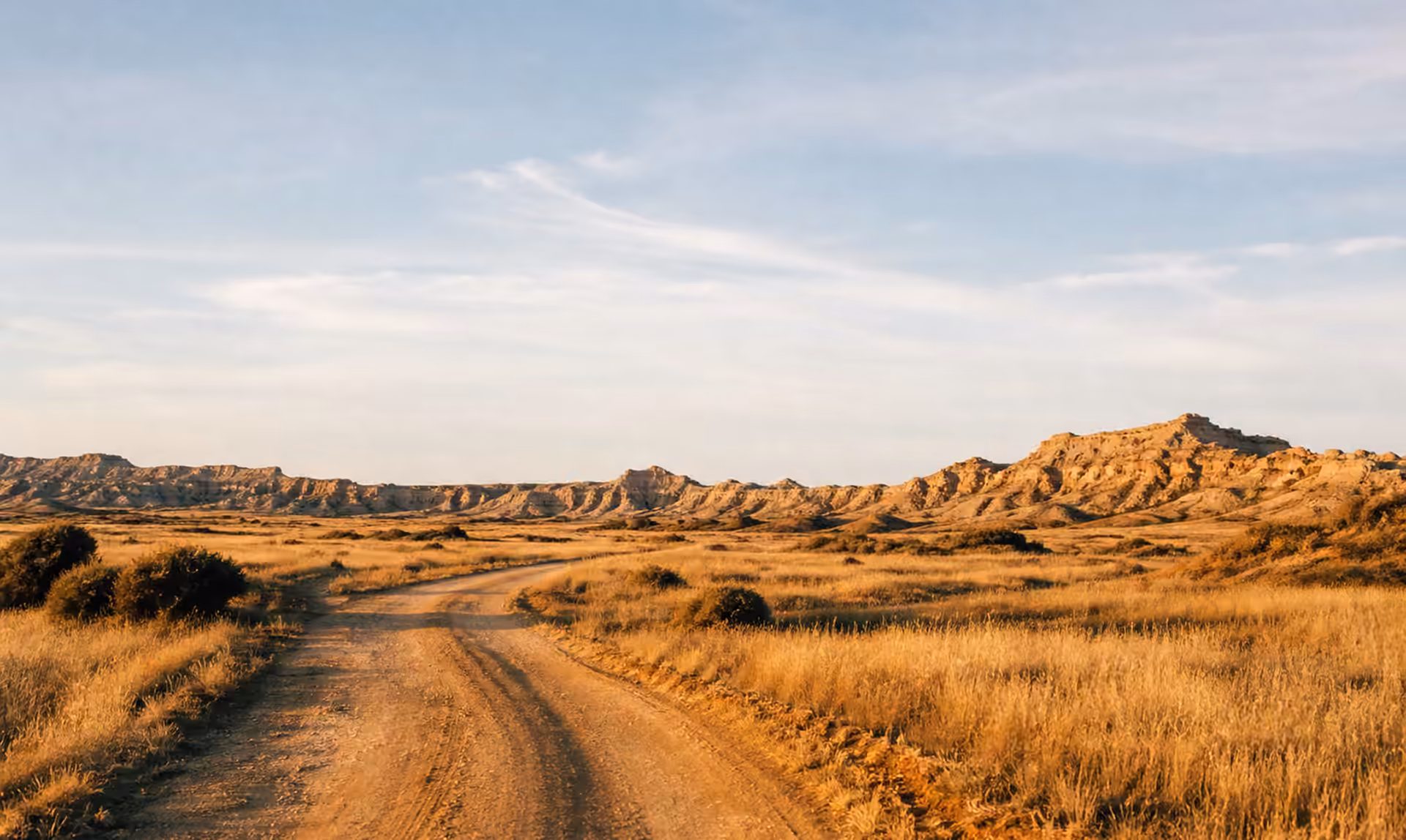 Dirt road through dry grass desert with rocky hills and blue sky.
