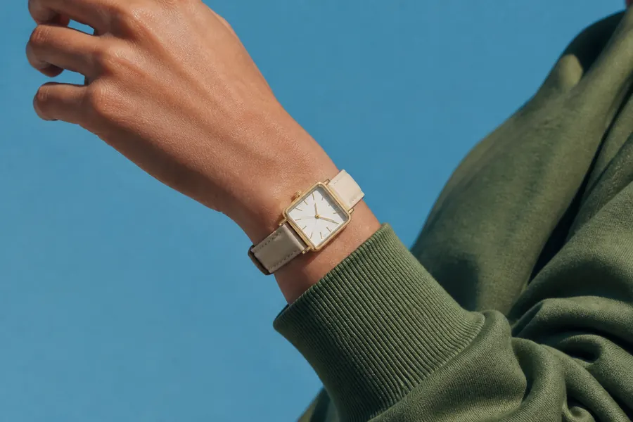A wrist wearing a square-faced watch against a bright sky