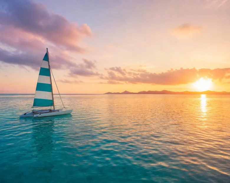 A small catamaran with a striped sail crossing a calm lagoon at sunset