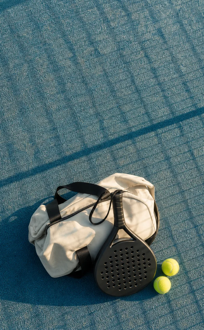 A canvas kit bag, a racket and two balls on a blue court crossed by fence shadows.